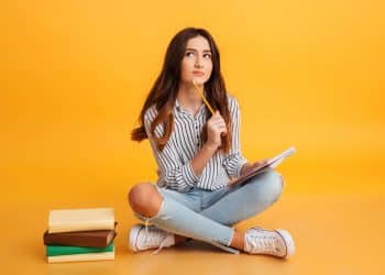 Estudante pensativa com caderno e lápis com fundo laranja.
