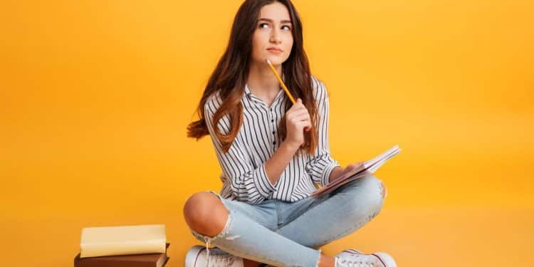Estudante pensativa com caderno e lápis com fundo laranja.