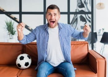 Torcedor vibrando com gol em casa no sofá laranja.