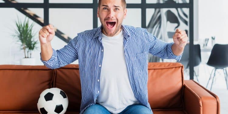 Torcedor vibrando com gol em casa no sofá laranja.