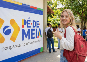Jovem loira com mochila vermelha faz sinal positivo ao lado de banner do programa Pé-de-Meia em pátio escolar.