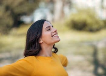 Mulher jovem de cabelos castanhos e moletom amarelo sorri com olhos fechados e braços abertos em parque.