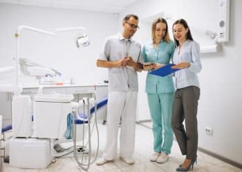 Dentista, assistente de uniforme azul-turquesa e mulher de camisa azul-clara analisam prancheta em consultório odontológico, simbolizando o novo piso salarial de médicos e cirurgiões-dentistas.