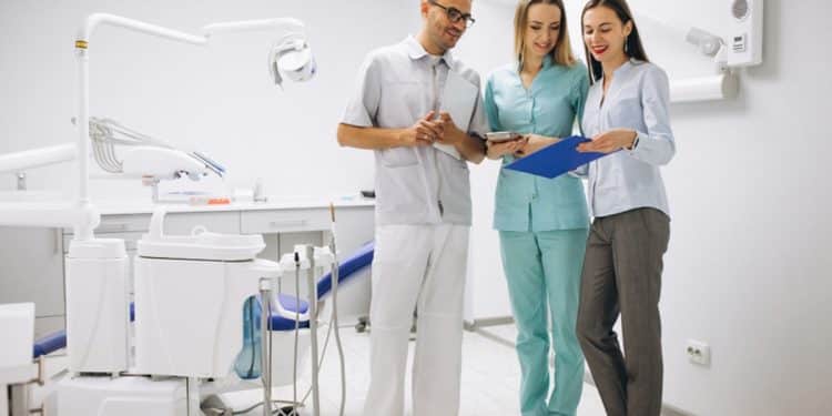 Dentista, assistente de uniforme azul-turquesa e mulher de camisa azul-clara analisam prancheta em consultório odontológico, simbolizando o novo piso salarial de médicos e cirurgiões-dentistas.