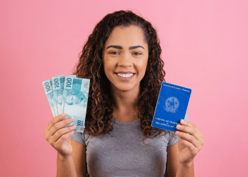 Jovem sorridente segura notas de 100 reais e Carteira de Trabalho e Previdência Social azul em fundo rosa.