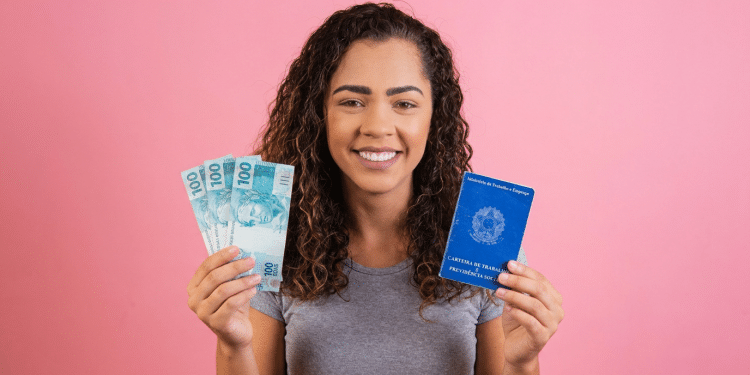 Jovem sorridente segura notas de 100 reais e Carteira de Trabalho e Previdência Social azul em fundo rosa.