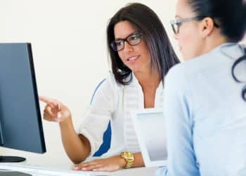 Duas mulheres em escritório, uma de blusa branca aponta para monitor enquanto outra de camisa azul observa atentamente, simbolizando a diferença entre RH e Departamento pessoal.