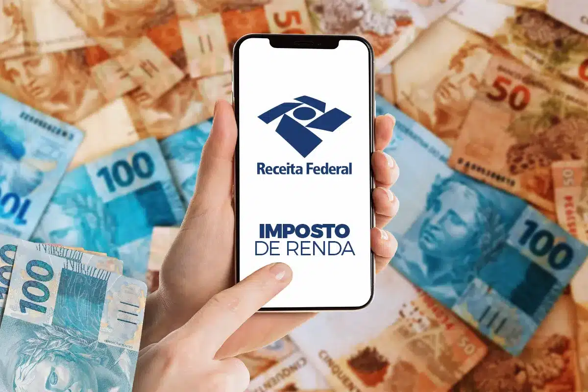 Mão segurando smartphone com tela da Receita Federal sobre cédulas brasileiras de 50 e 100 reais.