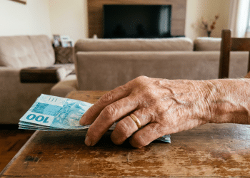 Mão de idosa segurando cédulas de R$ 100 em sala residencial.