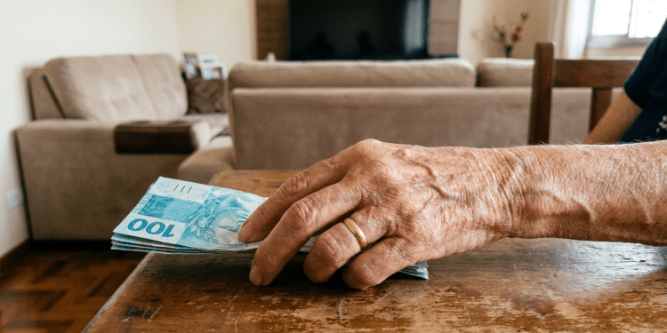 Mão de idosa segurando cédulas de R$ 100 em sala residencial.