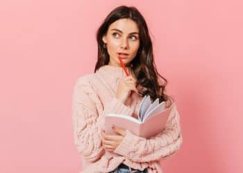 Mulher jovem de suéter rosa tricotado segura caderno e lápis vermelho enquanto olha pensativa para o lado, simbolizando a dúvida clássica entre " a gente vai" e " a gente vamos".