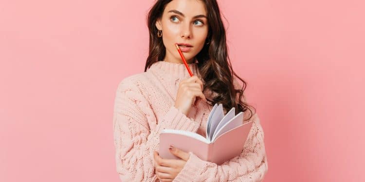 Mulher jovem de suéter rosa tricotado segura caderno e lápis vermelho enquanto olha pensativa para o lado, simbolizando a dúvida clássica entre " a gente vai" e " a gente vamos".