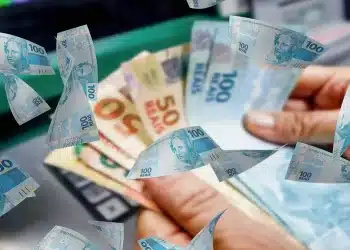 Notas de real sendo seguradas com outras cédulas de dinheiro ao fundo, representando o pagamento do Abono Salarial PIS/Pasep 2026 e a variação dos valores recebidos pelos trabalhadores.