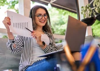 Mulher de óculos e fones de ouvido sorrindo segurando gráfico impresso enquanto trabalha no notebook em ambiente externo com plantas ao fundo