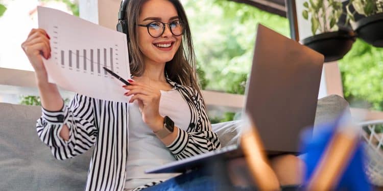 Mulher de óculos e fones de ouvido sorrindo segurando gráfico impresso enquanto trabalha no notebook em ambiente externo com plantas ao fundo