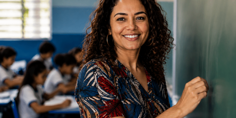 Professora sorri ao lado do quadro em uma sala de aula com alunos ao fundo