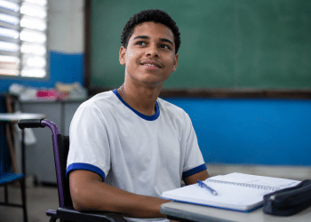 Jovem cadeirante com camiseta branca de gola azul sorri em sala de aula com lousa verde ao fundo.