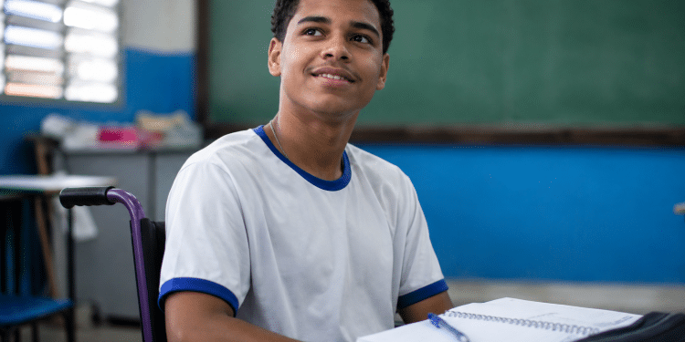 Jovem cadeirante com camiseta branca de gola azul sorri em sala de aula com lousa verde ao fundo.