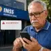 Idoso consultando aviso importante do INSS pelo celular em frente a prédio da Previdência Social, representando o bloqueio de pagamento para quem não atende à notificação.