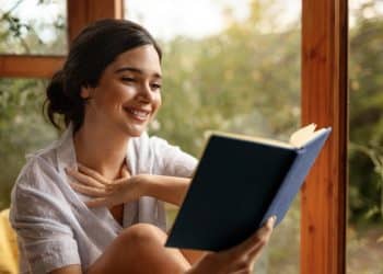 Mulher sorridente lendo livro azul próxima a uma janela com vegetação ao fundo