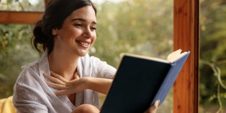 Mulher sorridente lendo livro azul próxima a uma janela com vegetação ao fundo