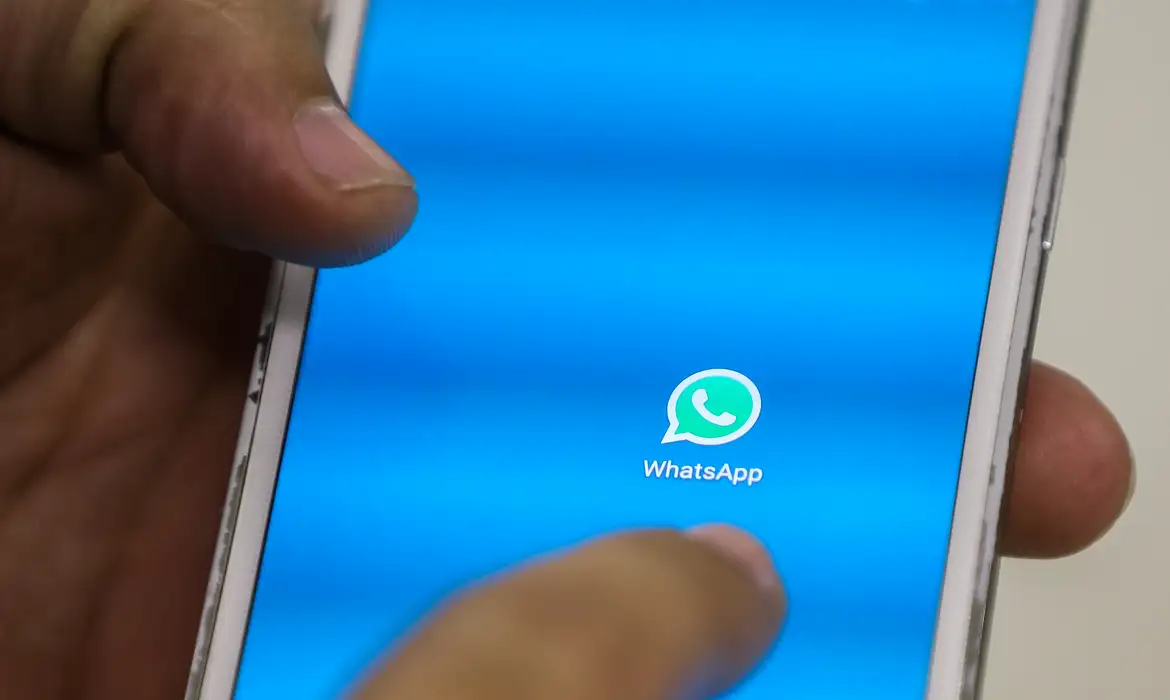 Pessoa segura o celular com o ícone do WhatsApp na tela, em referência ao bloqueio de contatos no aplicativo.