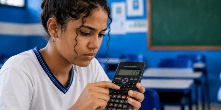 Jovem estudante com uniforme branco e azul concentrada usando calculadora científica em sala de aula com lousa verde.