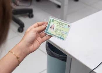 Mulher segura a nova Carteira de Identidade Nacional plastificada sobre balcão de atendimento em ambiente de repartição pública