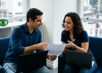 Duas pessoas discutem documento impresso com notebooks ao lado
