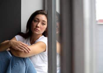 Jovem mulher de camiseta branca sentada junto à janela com expressão pensativa e olhar distante