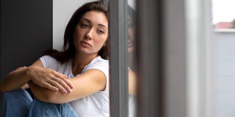 Jovem mulher de camiseta branca sentada junto à janela com expressão pensativa e olhar distante