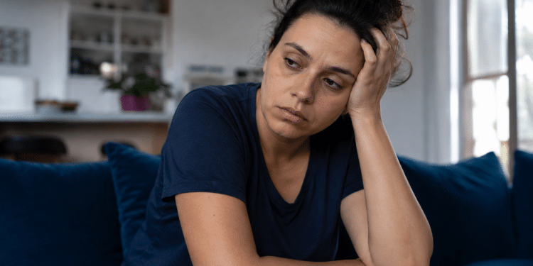 Mulher de camiseta azul-marinho sentada em sofá azul, apoiando a cabeça na mão com expressão cansada.