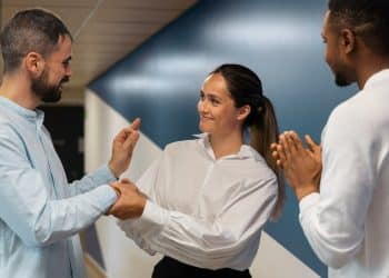 Mulher de camisa branca cumprimenta colega enquanto outro aplaude em ambiente corporativo com parede azul, simbolizando os comportamentos de quem teve muitos amigos na infância.