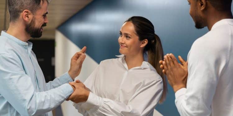 Mulher de camisa branca cumprimenta colega enquanto outro aplaude em ambiente corporativo com parede azul, simbolizando os comportamentos de quem teve muitos amigos na infância.