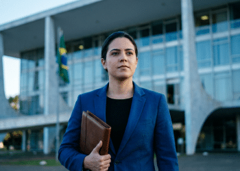 Mulher de blazer azul segura pasta marrom em frente ao Palácio do Planalto com bandeira do Brasil ao fundo.