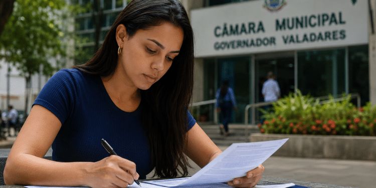 Mulher jovem com blusa azul-marinho preenche formulário em frente à Câmara Municipal de Governador Valadares.