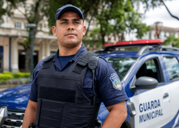 Guarda Municipal de uniforme azul marinho com colete balístico e rádio comunicador, diante de viatura oficial.