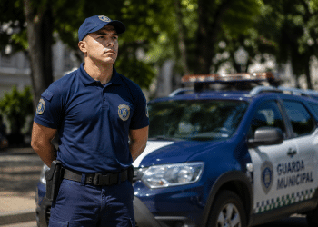 Agente da Guarda Municipal com uniforme azul-marinho e boné ao lado de viatura com inscrição "Guarda Municipal".