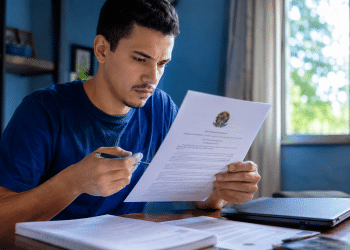 Homem de camiseta azul lê documento oficial com Brasão da República, segurando caneta, em home office com laptop.