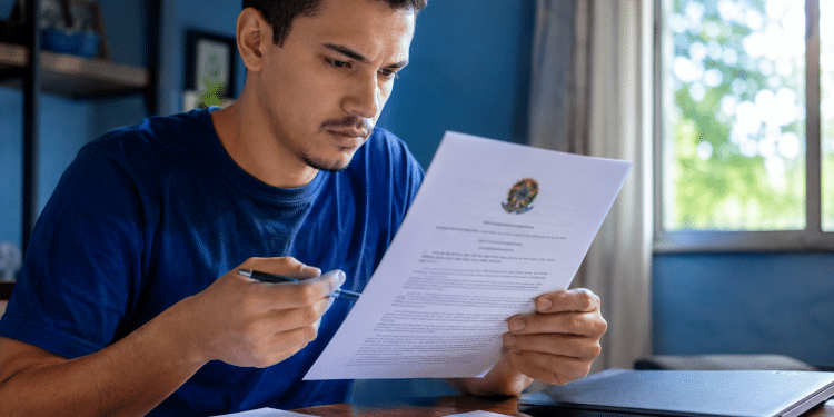 Homem de camiseta azul lê documento oficial com Brasão da República, segurando caneta, em home office com laptop.