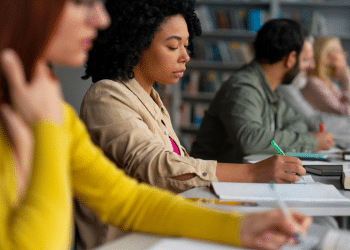 Jovem negra de cabelos cacheados escreve em caderno durante aula, com colegas ao fundo em biblioteca.