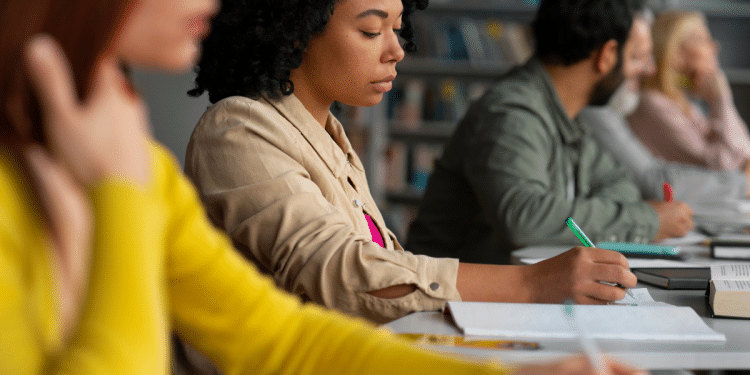 Jovem negra de cabelos cacheados escreve em caderno durante aula, com colegas ao fundo em biblioteca.