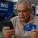 Aposentado do INSS consulta celular enquanto segura cartão CPF em agência da Previdência Social, representando o alerta sobre vazamento de dados de beneficiários em 2026.