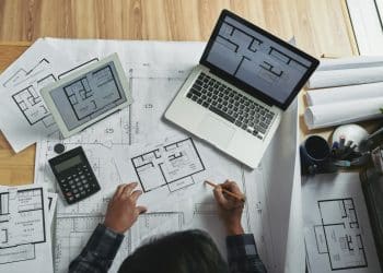 Mãos de arquiteto sobre plantas baixas residenciais com laptop e tablet exibindo projetos em mesa de madeira.