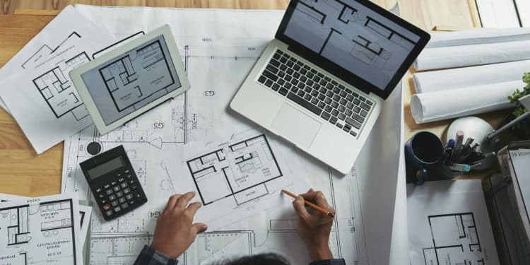 Mãos de arquiteto sobre plantas baixas residenciais com laptop e tablet exibindo projetos em mesa de madeira.