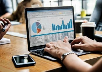 Profissional digita em laptop exibindo planilha Excel com gráfico pizza e barras, mesa de reunião com café, simbolizando a importância do curso Excel para a carreira.