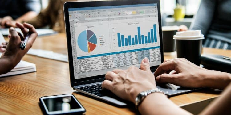 Profissional digita em laptop exibindo planilha Excel com gráfico pizza e barras, mesa de reunião com café, simbolizando a importância do curso Excel para a carreira.