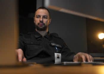 Profissional de segurança patrimonial com uniforme preto e crachá, operando computadores em central de monitoramento, simbolizando o curso de Segurança Patrimonial.