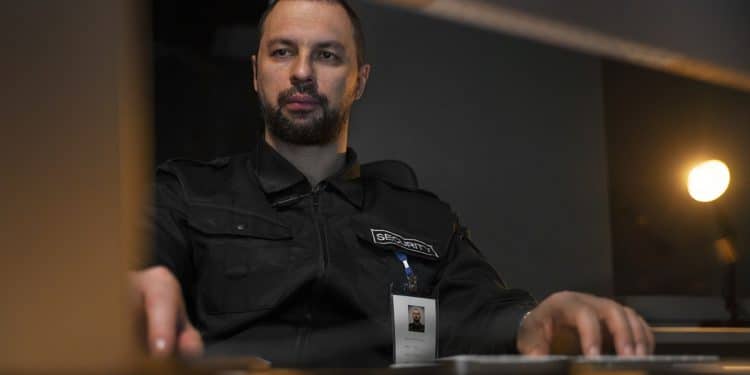 Profissional de segurança patrimonial com uniforme preto e crachá, operando computadores em central de monitoramento, simbolizando o curso de Segurança Patrimonial.