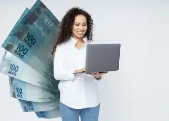 Profissional sorridente trabalhando com notebook ao lado de cédulas de R$ 100, em referência aos altos salários da carreira de desenvolvedor de inteligência artificial em regime de trabalho remoto.